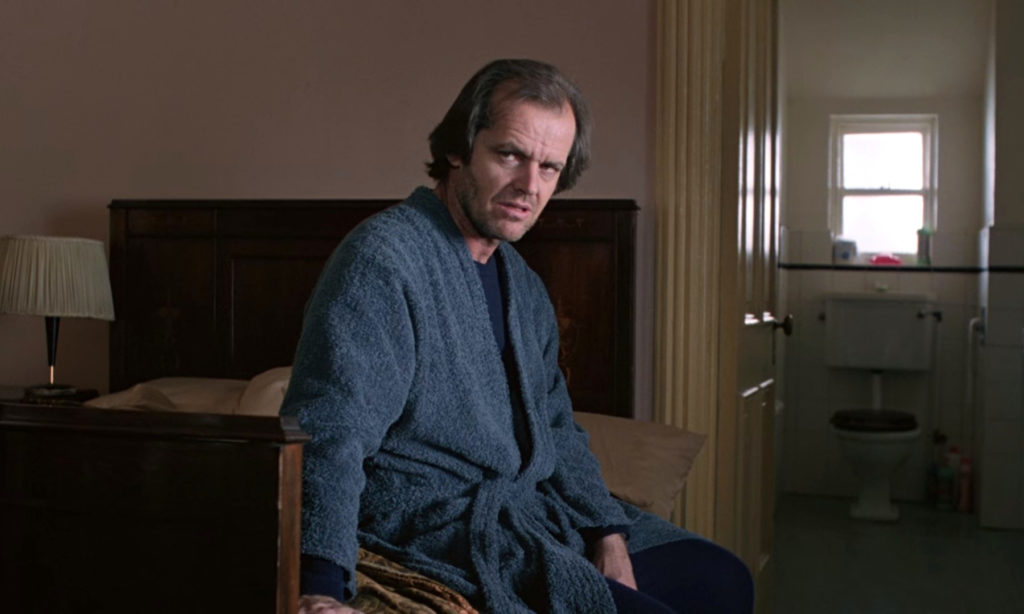Kadr z filmu "Lśnienie". Mężczyzna siedzi na łóżku ubrany w szary szlafrok. Jest to Jack Torrance, grany przez Jacka Nicholsona. W tle widoczna łazienka.