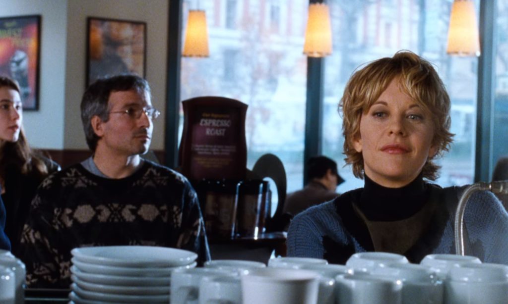 Scena z filmu "Masz wiadomość". Widok kawiarni Starbucks. Na pierwszym planie rząd białych filiżanek do kawy. Za nimi stoi zamyślona, lekko uśmiechająca się kobieta. Jest nią Katheelyn Kelly grana przez Meg Ryan, główna bohaterka. Za nią mężczyzna i kobieta czekający w kolejce.