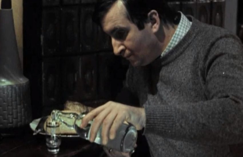 Scena z filmu brunet wieczorową porą. Michał Roman nalewa wódkę do kieliszka.
